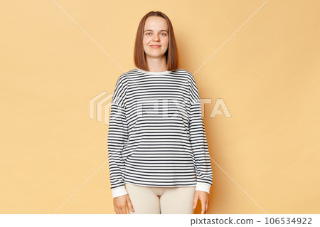 Smiling brown haired woman wearing casual clothing standing isolated over beige background looking at camera with positive facial expression. 106534922