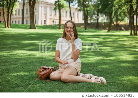 Positive Caucasian girl talking on cellphone while relaxing on in park, enjoying sunny day outdoors, wearing casdual clothing having free time enjoying summer weather. 106534923
