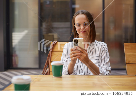Adorable positive smiling businesswoman in formal attire while using smartphones drinking coffee in cafe browsing internet checking socila networks while having break. 106534935