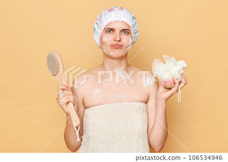 Sad upset disappointed woman wearing shower cap standing isolated over beige background holding washcloth and brush for dry massage. 106534946