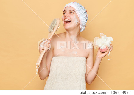 Happy funny woman wearing shower cap standing isolated over beige background being in bath posing wrapped in towel holding dry massage brush as microphone and washcloth in hand. Happy funny woman wearing shower cap standing isolated over beige background being in bath posing wrapped in towel holding dry massage brush as microphone and washcloth in hand. 106534951