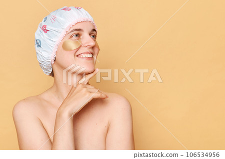 Attractive smiling woman wearing shower cap standing isolated over beige background using patched for under eye skin care looking away with dreaming expression. Attractive smiling woman wearing shower cap standing isolated over beige background using patched for under eye skin care looking away with dreaming expression. 106534956