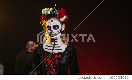 Holy goddess of death posing in halloween costume with skull make up to celebrate dios de los muertos on mexican holiday. Spooky woman as santa muerte with black and white body art. Handheld shot. Holy goddess of death posing in halloween costume with skull make up to celebrate dios de los muertos on mexican holiday. Spooky woman as santa muerte with black and white body art. Handheld shot. 106535012