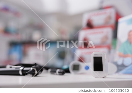 Medical oximeter standing on table in empty drugstore ready to be used by client, digital device to measure oxygen saturation. Pharmacy filled with medicaments, vitamins and pharmaceutical products 106535020