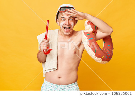 Happy overjoyed tourist man at summer resort standing with rubber ring isolated over yellow background looking far with hand near forehead laughing happily. 106535061