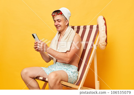 Smiling happy attractive young tourist man in summer clothes and panama sitting on deck chair isolated over yellow background holding samrtphone browsing internet at resort. 106535079