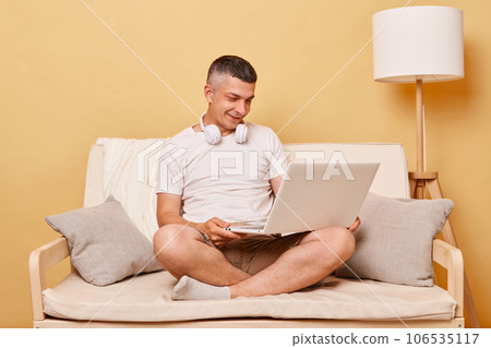 Pleased positive man wearing white T-shirt and shorts sitting on sofa against beige background looking at laptop display watching movie working from home online. 106535117