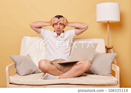 Tired exhausted man wearing white T-shirt and shorts sitting on sofa against beige background having headache tribles with work deadline holding computer. Tired exhausted man wearing white T-shirt and shorts sitting on sofa against beige background having headache tribles with work deadline holding computer. 106535119