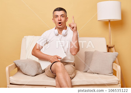 Inspired excited man wearing white T-shirt and shorts sitting on sofa against beige background working on laptop looking at camera raised finger up having idea. 106535122