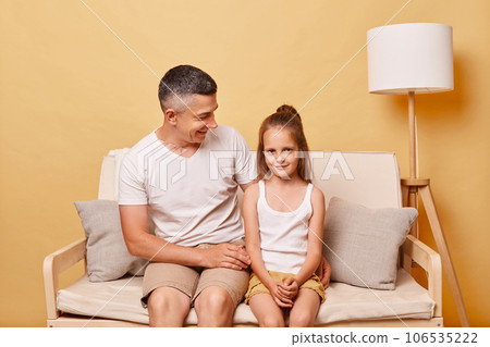 Positive handsome man father with child daughter wearing casual clothing sitting on sofa against beige background dad talking worth his female child. 106535222