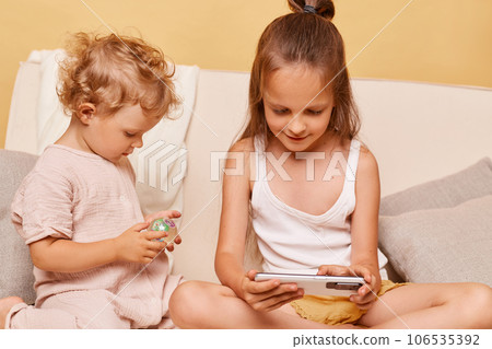 Closeup portrait of charming curious little girls sisters using phone while sitting on sofa at home against beige wall using smartphone browsing internet kids content. 106535392