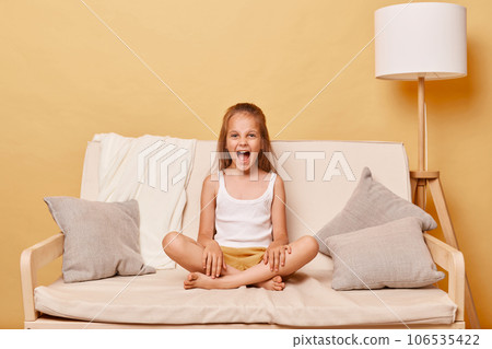 Angry little girl wearing casual clothing sitting on sofa at home against beige wall having problems during teenage years having hysterical behavior screaming. 106535422