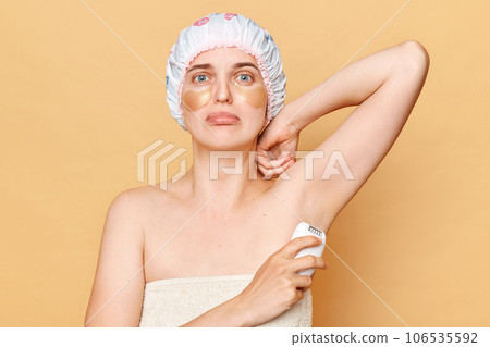 Unhappy woman wearing shower cap standing isolated over beige background posing with patches under eyes shaving her armpit taking care of her skin. 106535592