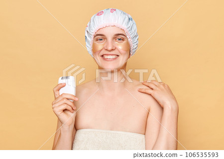 Optimistic woman wearing shower cap standing isolated over beige background holding depilator smiling happily posing in towel morning routine. 106535593