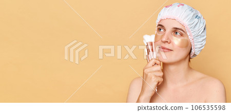 Pensive woman wearing shower cap standing isolated over beige background looking away at copy space for promotion holding makeup brushes morning beauty routine Pensive woman wearing shower cap standing isolated over beige background looking away at copy space for promotion holding makeup brushes morning beauty routine 106535598