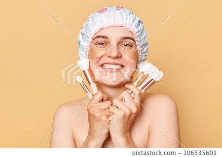 Joyful woman wearing shower cap standing isolated over beige background posing with big variety of makeup brushes doing beauty routine procedures after taking shower. Joyful woman wearing shower cap standing isolated over beige background posing with big variety of makeup brushes doing beauty routine procedures after taking shower. 106535601