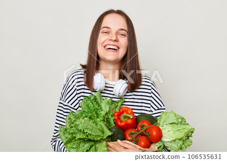 Enjoying a vibrant green diet. Fresh greenery and vegetables. Happy laughing woman wearing striped casual shirt carrying lettuce cabbage pepper avocado isolated over gray background 106535631