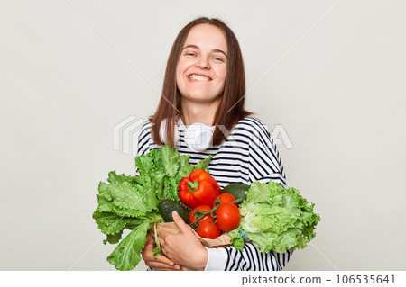 Satisfied delighted woman wearing striped casual shirt carrying vegetable isolated over gray background looking at camera with toothy smile laughing. Satisfied delighted woman wearing striped casual shirt carrying vegetable isolated over gray background looking at camera with toothy smile laughing. 106535641