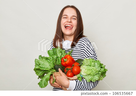 Satisfied woman wearing striped casual shirt holding lettuce cabbage pepper avocado isolated over gray background showing tongue out enjoying healthy nutrition. Satisfied woman wearing striped casual shirt holding lettuce cabbage pepper avocado isolated over gray background showing tongue out enjoying healthy nutrition. 106535667