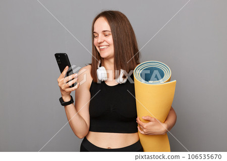 Smiling happy woman holding rolled fitness mat dressed in activewear isolated over gray background using modern mobile phone downloads application for sport training. Smiling happy woman holding rolled fitness mat dressed in activewear isolated over gray background using modern mobile phone downloads application for sport training. 106535670