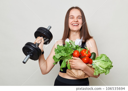 Extremely happy woman wearing sportswear carrying vegetable and barbel isolated over gray background doing physical exercising looking smiling at camera. 106535694