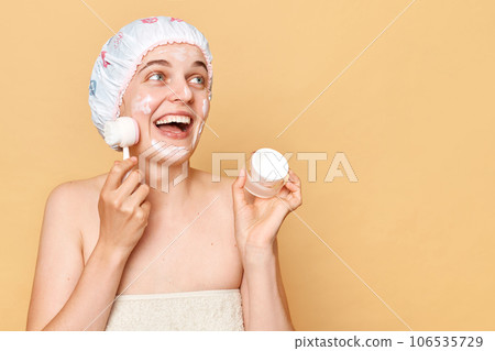 Excited attractive woman wearing shower cap standing isolated over beige background looking away at copy space for advertisement making cleansing procedures for her face. Excited attractive woman wearing shower cap standing isolated over beige background looking away at copy space for advertisement making cleansing procedures for her face. 106535729