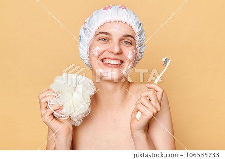 Attractive smiling woman woman wearing shower cap standing isolated over beige background holding toothbrush posing after taking shower. Attractive smiling woman woman wearing shower cap standing isolated over beige background holding toothbrush posing after taking shower. 106535733