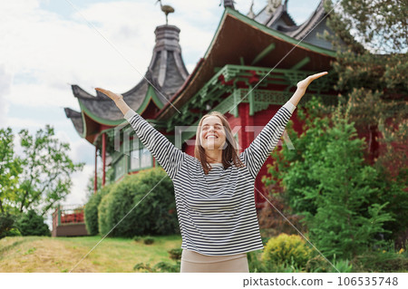 Carefree happy woman in spring or summer park raising arms with happiness, caucasian girl relaxing and enjoying life on nature outdoors posing with beautiful building on background. Carefree happy woman in spring or summer park raising arms with happiness, caucasian girl relaxing and enjoying life on nature outdoors posing with beautiful building on background. 106535748