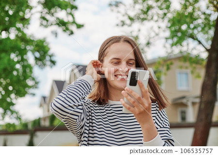 Woman in town browsing internet on smart phone making selfie having video call smiling happily browsing internet outdoor touching her hair wearing striped shirt. Woman in town browsing internet on smart phone making selfie having video call smiling happily browsing internet outdoor touching her hair wearing striped shirt. 106535757