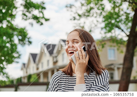 happy Caucasian young woman talking with cellphone in street with buildings on background, holding mobile phone laughing having pleasant conversation with friends. happy Caucasian young woman talking with cellphone in street with buildings on background, holding mobile phone laughing having pleasant conversation with friends. 106535758
