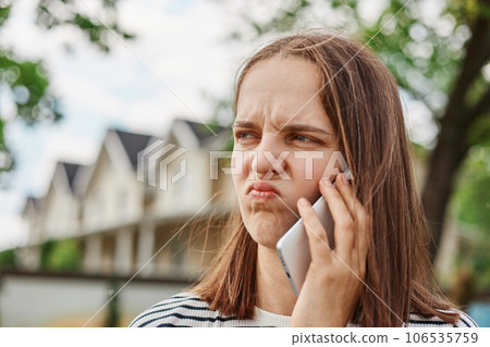 Unhappy sad brown haired young woman talking with cellphone frowning face expressing negative emotions standing outdoor with houses on background. Unhappy sad brown haired young woman talking with cellphone frowning face expressing negative emotions standing outdoor with houses on background. 106535759