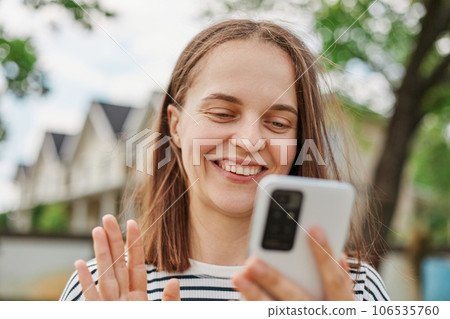 Distant Communication. Young adult woman with smartphone making video call outdoors, smiling female waving hand at cellphone camera while walking on street. Distant Communication. Young adult woman with smartphone making video call outdoors, smiling female waving hand at cellphone camera while walking on street. 106535760
