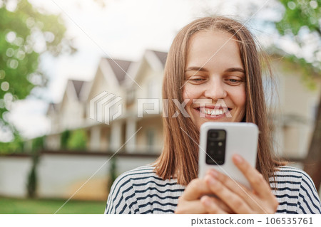 Smiling brown haired woman browsing internet on smart phone standing outdoor in street with houses on background checking social network while walking outside. Smiling brown haired woman browsing internet on smart phone standing outdoor in street with houses on background checking social network while walking outside. 106535761