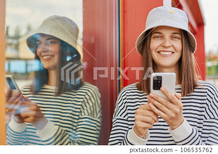 Young beautiful woman wearing striped shirt and panama using app in her smartphone device sending text message standing near building with big window reflection. 106535762