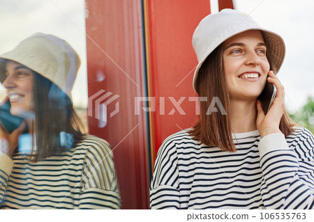 Portrait of smiling woman wearing striped shirt and panama talking with cellphone outdoors standing near building with window looking away copy space. 106535763