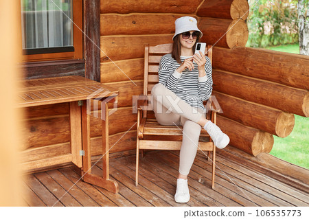 Positive woman wearing striped shirt panama sunglasses sitting on terrace of wooden house using mobile phone chatting online with friends browsing internet. 106535773