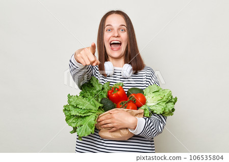 Healthy nutrition. Excited woman with brown hair wearing striped shirt standing isolated over gray background pointing at camera holding vegetable in hands. Healthy nutrition. Excited woman with brown hair wearing striped shirt standing isolated over gray background pointing at camera holding vegetable in hands. 106535804