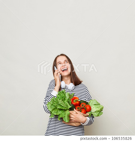 Amazed woman with brown hair wearing striped shirt standing isolated over gray background talking on mobile phone looking up at advertisement area holding vegetables Amazed woman with brown hair wearing striped shirt standing isolated over gray background talking on mobile phone looking up at advertisement area holding vegetables 106535826