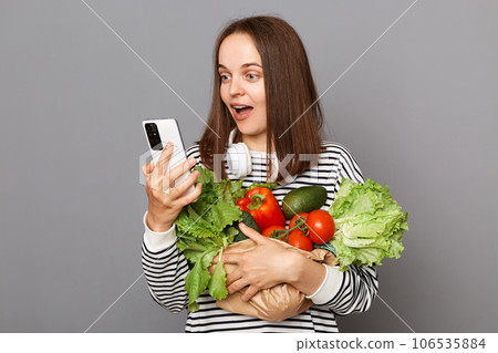 Amazed woman holding vegetables isolated over gray background holding mobile phone using internet searching information about healthy eating. 106535884