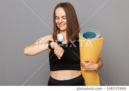 Smiling young girl wearing black top holding fitness mat in her hands posing isolated over gray background checking burned calories on her fitness tracker. Smiling young girl wearing black top holding fitness mat in her hands posing isolated over gray background checking burned calories on her fitness tracker. 106535969