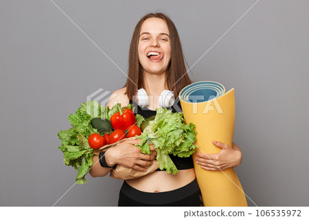Delighted Caucasian woman holding organic vegetables and karemat standing isolated over gray background showing tongue being hungry after hard sport training. 106535972