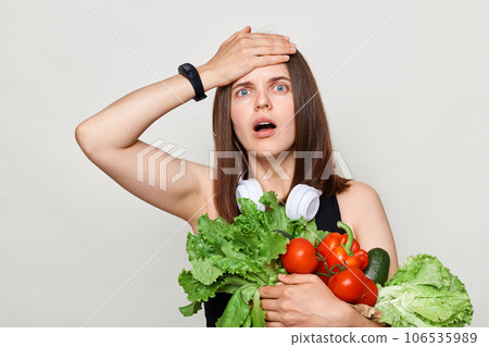 Shocked adult woman with brown hair standing isolated over white background holding vegetables making facepalm gesture looking at camera with big eyes. 106535989
