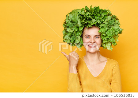 Fresh food care. Healthy eating habits. woman with lettuce hair style with green vegetables on her head isolated over yellow background pointing at advertisement area, copy space. Fresh food care. Healthy eating habits. woman with lettuce hair style with green vegetables on her head isolated over yellow background pointing at advertisement area, copy space. 106536033