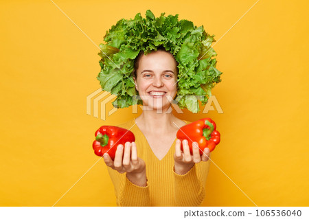 Friendly woman wearing casual shirt and lettuce wreath holding bell pepper standing isolated over yellow background offering fresh bell peppers looking at camera. 106536040
