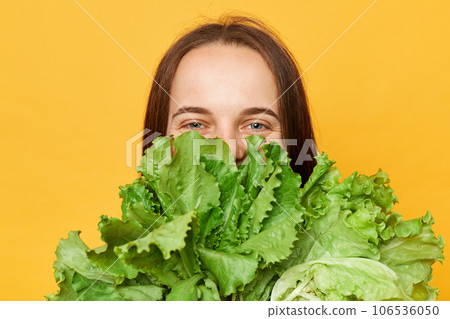 Weight loss goals. Slimming meal plan. Green and nutritious. Cheerful woman covering half of face with lettuce green vegetables isolated over yellow background looking at camera. 106536050