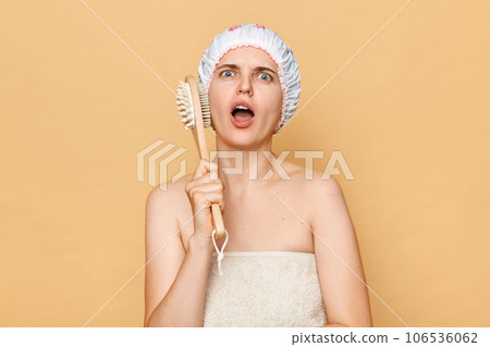 Shocked scared caucasian woman with massage brush on beige background taking shower, looking at camera with open mouth and displeased expression. 106536062