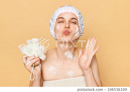 Relaxation self-care routine. Healthy hygiene habits. Positive optimistic young woman in shower cap and wrapped in towel sending air kissing isolated over beige background 106536109