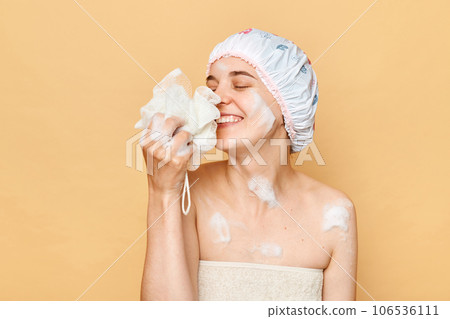 Smiling delighted woman with foam on her face and body wearing bath cap and towel standing with washcloth smelling pleasant aromat of her shower gel enjoying. 106536111