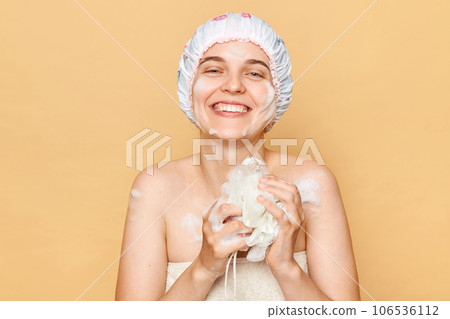 Satisfied positive smiling female dressed bath cap looking at camera with toothy smile holding sponge with foam taking shower in thew morning expressing happiness. 106536112