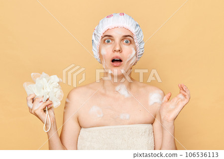 Shocked astonished surprised female dressed bath cap looking at camera with open mouth holding sponge with foam taking shower in thew morning expressing fear. Shocked astonished surprised female dressed bath cap looking at camera with open mouth holding sponge with foam taking shower in thew morning expressing fear. 106536113